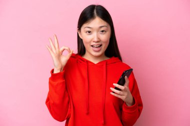 Pembe arka planda izole edilmiş genç Çinli bir kadın cep telefonu kullanıyor ve tamam işareti yapıyor.