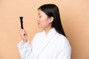 Bej arka planda izole edilmiş genç Çinli kadın elinde makyaj fırçası tutuyor.