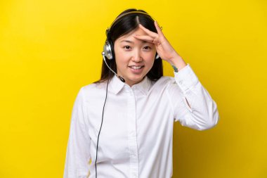 Tele pazarlamacı Çinli kadın sarı arka planda izole edilmiş bir kulaklıkla çalışıyor. Bir şeye bakmak için çok uzaklara bakıyor.