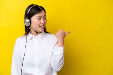 Sarı arka planda izole edilmiş kulaklıkla çalışan pazarlamacı Çinli kadın bir ürünü sunmak için yan tarafı işaret ediyor.