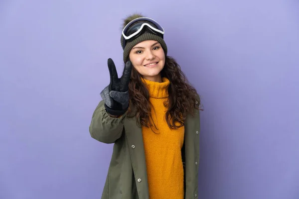 俄罗斯少女 戴着雪板眼镜 与紫色背景隔离 面带微笑 显示出胜利的迹象 — 图库照片