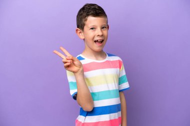 Mor arka planda izole edilmiş küçük çocuk gülümsüyor ve zafer işareti gösteriyor.