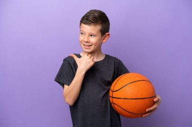 Little boy playing basketball isolated on purple background pointing to the side to present a product
