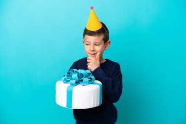 Mavi arka planda izole edilmiş doğum günü pastası tutan küçük çocuk yana bakıyor ve gülümsüyor.