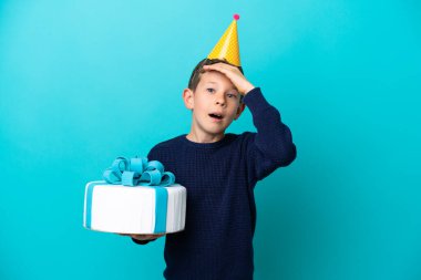 Mavi arka planda izole edilmiş doğum günü pastası tutan küçük çocuk yan tarafa bakarken sürpriz jest yapıyor.