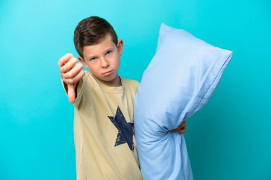 Mavi arka planda izole edilmiş pijamalı küçük bir çocuk negatif ifadeyle baş parmağını aşağı gösteriyor.