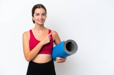 Beyaz arka planda izole edilmiş bir minder tutarak yoga derslerine giden beyaz bir kadın bir ürünü sunmak için yan tarafı işaret ediyor.