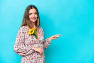 Ayçiçeği tutan beyaz bir kadın. Mavi arka planda izole edilmiş. Parmağını yan tarafa gösteriyor.