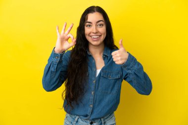 Sarı arka planda izole edilmiş genç, beyaz bir kadın tamam işareti ve baş parmak hareketi gösteriyor.