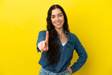 Sarı arka planda izole edilmiş genç beyaz kadın parmağını kaldırıyor ve gösteriyor.