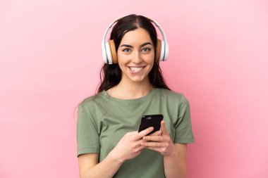 Pembe arka planda izole edilmiş genç beyaz bir kadın cep telefonuyla müzik dinliyor ve ön tarafa bakıyor.