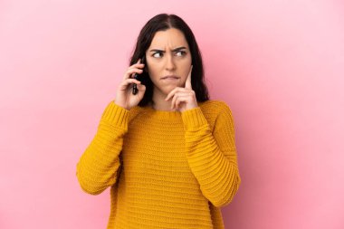 Pembe arka planda izole edilmiş cep telefonu kullanan beyaz bir kadın. Şüpheleri ve düşünceleri var.