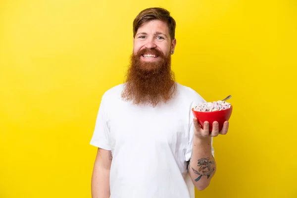 Redhead man with beard eating a bowl of cereals isolated on yellow background smiling a lot