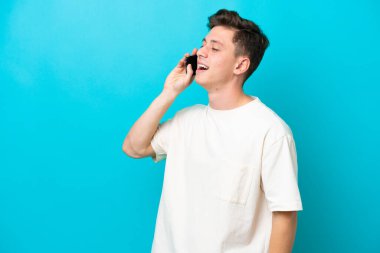 Genç yakışıklı Brezilyalı adam mavi arka planda izole edilmiş cep telefonuyla sohbet ediyor.