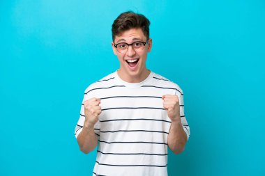 Genç yakışıklı Brezilyalı adam mavi arka planda izole edilmiş galibiyeti kutluyor.
