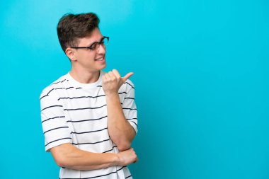 Mavi arka planda izole edilmiş yakışıklı Brezilyalı genç bir adam bir ürün sunmak için kenarı işaret ediyor.