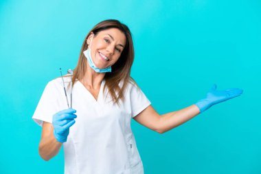 Mavi arka planda izole edilmiş orta yaşlı doktor kadın davet etmek için ellerini yana uzatıyor.