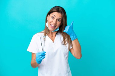 Mavi arka planda izole edilmiş orta yaşlı bir doktor telefon jesti yapıyor. Bana geri dönüş işareti ver.