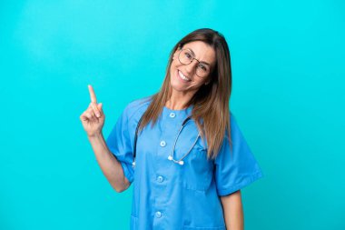 Mavi arka planda izole edilmiş orta yaşlı cerrah kadın en iyinin işareti olarak parmağını kaldırıyor ve gösteriyor.