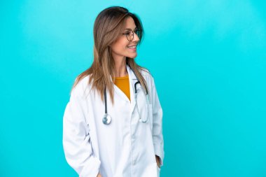 Mavi arka planda izole edilmiş orta yaşlı kadın doktor önlüğü giyiyor ve steteskop kullanıyor.