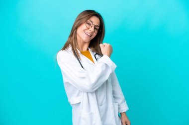 Mavi arka planda soyutlanmış orta yaşlı bir kadın doktor önlüğü giyiyor ve zafer jesti yapıyor.