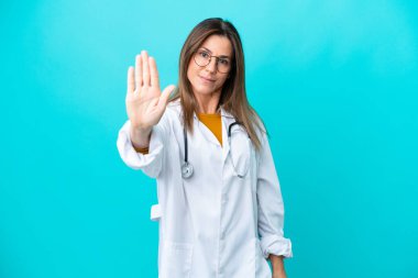 Mavi arka planda izole edilmiş orta yaşlı bir kadın doktor önlüğü giyiyor ve dur işareti yapıyor.