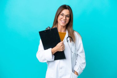 Mavi arka planda izole edilmiş orta yaşlı bir kadın doktor önlüğü giyiyor ve elinde bir klasör tutuyor.