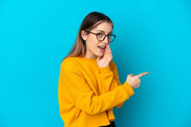 Mavi arka planda izole edilmiş genç beyaz kadın bir ürünü sunmak ve bir şeyler fısıldamak için kenarı işaret ediyor.