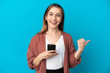 Genç beyaz kadın mavi arka planda izole edilmiş cep telefonu kullanıyor ve yan tarafı işaret ediyor.