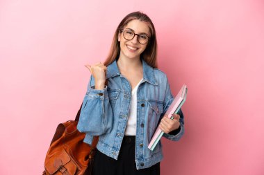 Pembe arka planda izole edilmiş genç beyaz kadın bir ürünü sunmak için kenarı işaret ediyor.