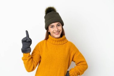 Kışlık şapkalı genç kız beyaz arka planda izole edilmiş ve en iyinin işareti olarak parmağını kaldırıyor.