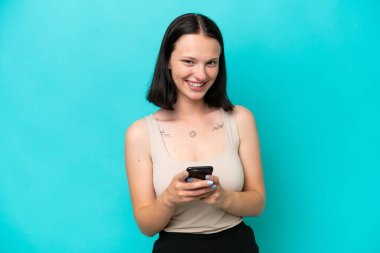 Mavi arka planda izole edilmiş genç beyaz kadın cep telefonuyla bir mesaj gönderiyor.