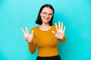 Mavi arka planda izole edilmiş genç beyaz kadın parmaklarıyla sekiz sayıyor.