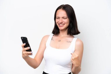 Beyaz arka planda izole edilmiş genç beyaz kadın cep telefonu kullanıyor ve zafer jesti yapıyor.