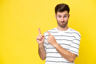 Sarı arka planda izole edilmiş genç beyaz adam korkmuş ve kenarı işaret ediyor.
