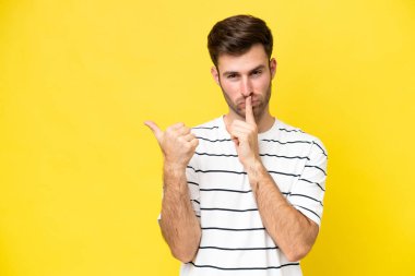 Sarı arka planda izole edilmiş genç beyaz bir adam yan tarafı işaret ediyor ve sessizlik hareketi yapıyor.