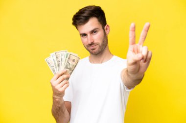Kafkasyalı genç adam sarı arka planda izole edilmiş bir sürü para alıyor gülümsüyor ve zafer işareti gösteriyor.