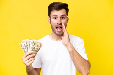 Kafkasyalı genç bir adam, sarı arka planda, şaşırmış ve şok olmuş bir yüz ifadesiyle izole edilmiş bir sürü para alıyor.