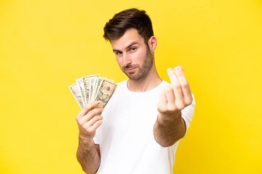Kafkasyalı genç adam çok para alıyor. Sarı arka planda izole edilmiş. Para hareketi yapıyor.