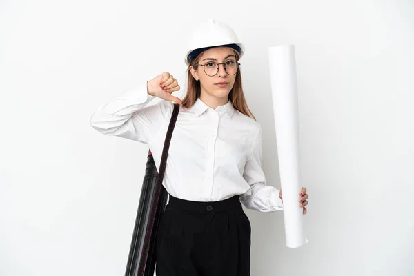 Young architect woman with helmet and holding blueprints isolated on white background showing thumb down with negative expression