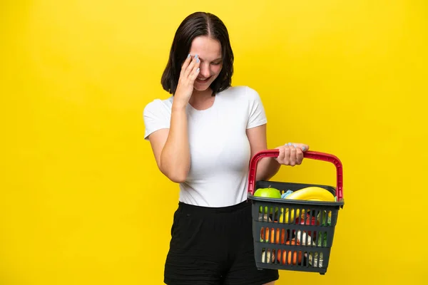 一个年轻的女人拿着一个购物篮 里面装满了被黄色背景的笑声隔开的食物 — 图库照片