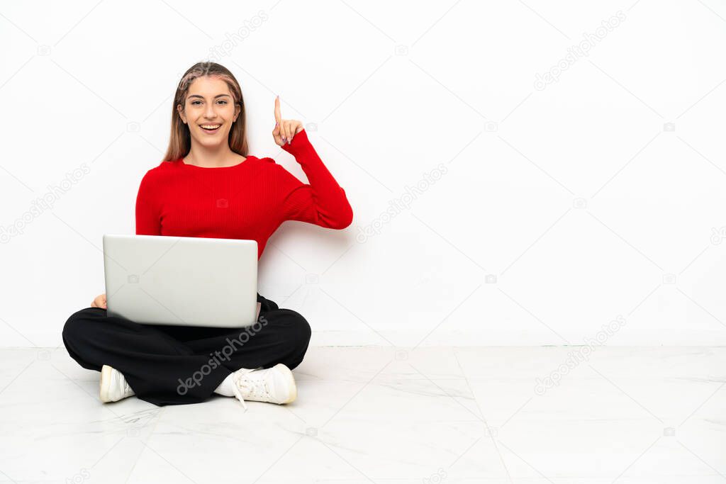 Young caucasian woman with a laptop sitting on the floor intending to realizes the solution while lifting a finger up