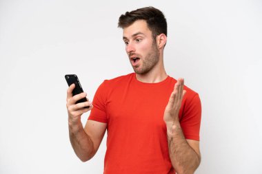 Beyaz arka planda izole edilmiş genç beyaz bir adam kameraya bakarken şaşırmış bir ifadeyle cep telefonunu kullanıyor.