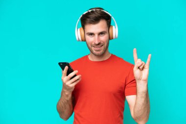 Kafkasyalı genç adam mavi arka planda izole edilmiş. Mobil bir rock hareketi yaparak müzik dinliyor.