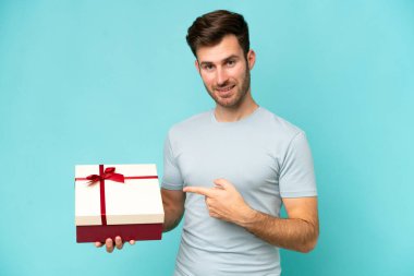 Young caucasian man holding a gift isolated on blue background and pointing it