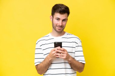 Sarı arka planda izole edilmiş genç beyaz adam cep telefonuyla bir mesaj gönderiyor.