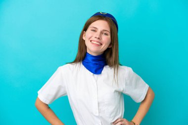 Uçak hostesi İngiliz kadın mavi arka planda izole edilmiş, kolları kalçasında ve gülümsüyor.