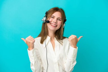 Tele pazarlamacı İngiliz kadın mavi arka planda izole edilmiş bir kulaklıkla çalışıyor. Baş parmakları havada ve gülümsüyor.