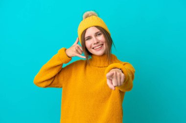 Kışlık ceket giyen genç İngiliz kadın mavi arka planda izole edilmiş telefon hareketi yapıyor ve ön tarafı işaret ediyor.