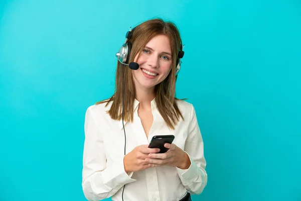 テレマーケティング担当者青い背景に隔離されたヘッドセットで働く英語の女性は 携帯電話でメッセージを送信します — ストック写真